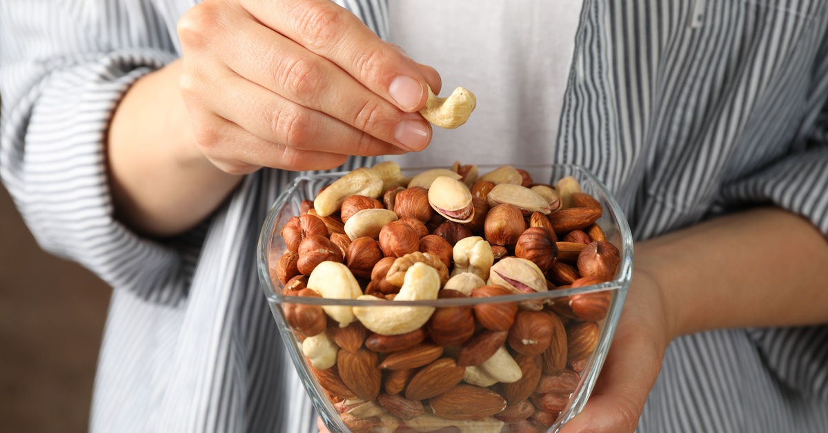 A person wearing a striped shirt holds a square glass bowl filled with mixed nuts and picks up a single cashew.