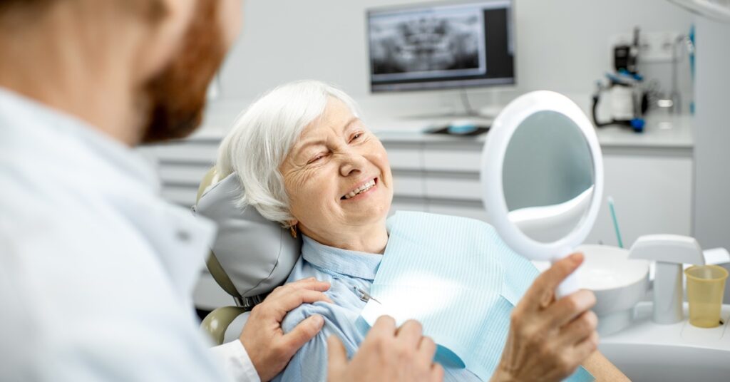 An older woman reclines in a dental chair and holds a mirror while smiling next to a dentist inside a clinic.