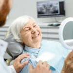 An older woman reclines in a dental chair and holds a mirror while smiling next to a dentist inside a clinic.