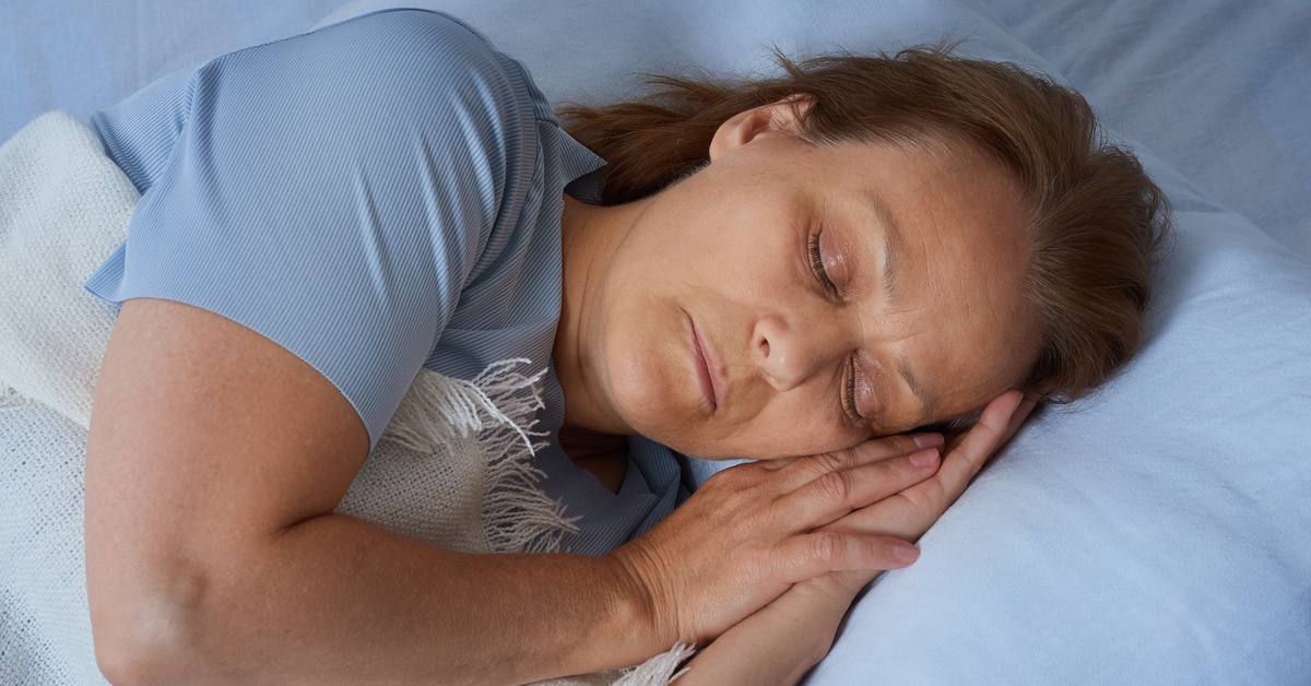 A woman lying on her side in bed with her hands together under her cheek and a white blanket over her torso.