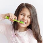 A young girl with long brown hair brushing her teeth using a green toothbrush while standing against a white wall.
