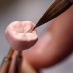 A close-up of a technician holding up a dental crown with tweezers and using a brush to apply ceramic material to the surface.