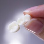 A hand holds a three-tooth dental bridge between its thumb and index finger against a softly blurred gray background.
