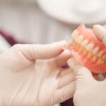 A person wearing gloves holding a full upper and lower denture set inside a clean, brightly lit dental room.